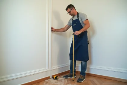 Artisan mesurant une plinthe haute contre un mur blanc dans une petite pièce avec parquet en bois